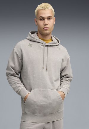 Sweatshirt à capuche gris clair en tissu doux, avec une poche avant et des accents de logo subtils. Le tissu a une finition légèrement texturée.