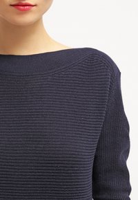 Pull tricot bleu marine avec un large col bateau et un côtes texturées. Comprend des manches longues et une silhouette ajustée.