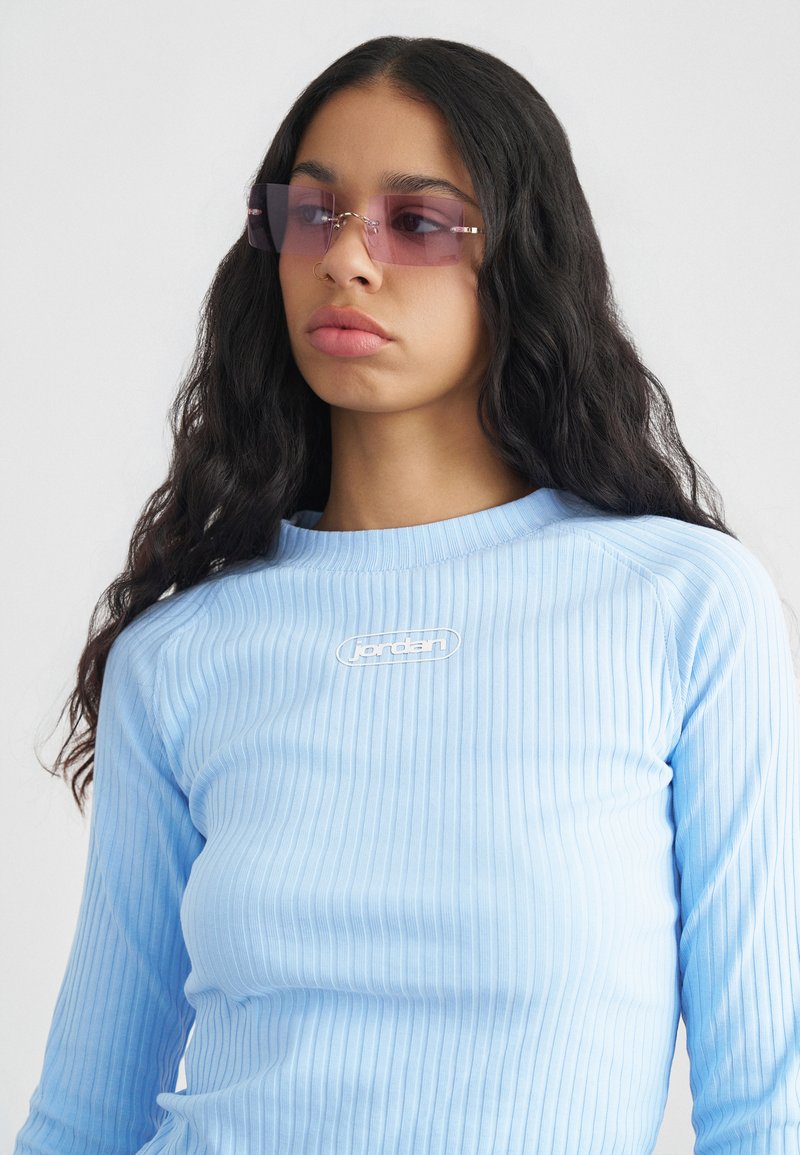 Νεαρή γυναίκα με μακριά, σγουρά μαλλιά που φοράει ορθογώνια ροζ-τυπωμένα γυαλιά ηλίου και μπλε ανοιχτού χρώματος μπλούζα με ριμπ και μακριά μανίκια με το λογότυπο "Jordan".