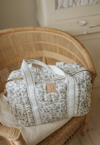 Quilted floral-patterned white and gray duffle bag with white handles on a wicker chair, next to a cream-colored cabinet.