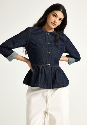 Nainen pukeutuneena tummaan farkkutakkiin, jossa on napitettu etuosa, kerätty vyötärö ja käärityt hihat, yhdistettynä väljäsäärisiin valkoisiin housuihin, poseeraa yksinkertaisen taustan edessä.