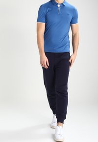 Modrá polokošile s krátkým rukávem s bílým límečkem, v kombinaci s námořnickými tepláky a bílými teniskami, vytvářející neformální vzhled.