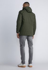 Chubasquero verde con capucha y una textura suave, combinado con unos jeans gris claro. El modelo lleva zapatillas blancas con suela de goma.