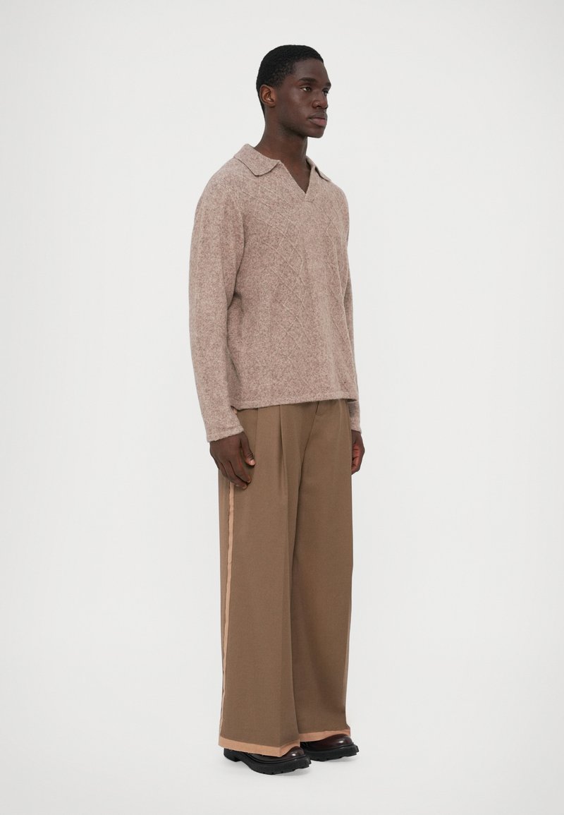 Pull en maille marron clair avec un col en V, associé à un pantalon large marron présentant une bande sur le côté et des chaussures sombres.