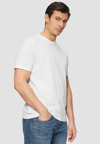 Junger Mann mit dunklem Haar, der ein schlichtes weißes T-Shirt und blaue Jeans trägt, steht mit einer Hand in seiner Tasche vor einem schlichten Hintergrund.