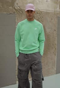 Sweatshirt verde menta com gola redonda e pequeno logotipo branco; combinado com calças cargo cinza-escuras com vários bolsos; boné de basebol rosa-claro.