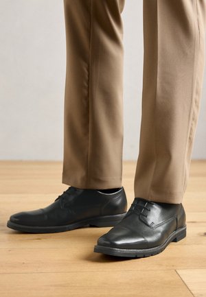 Zwarte leren dress shoes gedragen met beige broeken, staand op een lichte houten vloer.