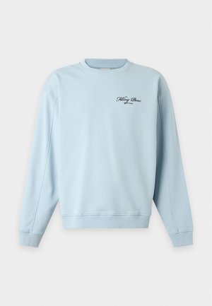 Homme portant un sweat-shirt bleu clair avec un petit texte, un short large noir jusqu'au genou, des chaussettes blanches et des baskets blanches, debout devant un fond uni.
