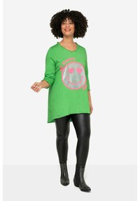 Grüne Tunika mit grafischem Silber- und Pink-Smiley, runder Ausschnitt und dreiviertel Ärmeln, kombiniert mit glänzenden schwarzen Leggings und beschlagenen Stiefeln.