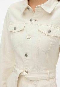 Gros plan sur une veste beige crème à boutons, avec des poches poitrine et une ceinture en tissu nouée à la taille.