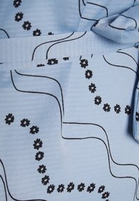 Un tessuto azzurro chiaro presenta un motivo testurizzato con linee ondulate nere e piccoli motivi floreali neri. Include un dettaglio in vita annodato.