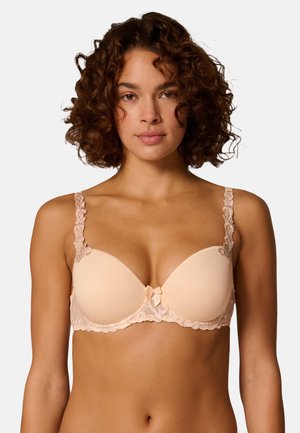 Frau mit lockigem Haar, die einen hellbeigen, gepolsterten BH mit Spitzendetails an Trägern und Band trägt, blickt nach vorne vor einem weißen Hintergrund.
