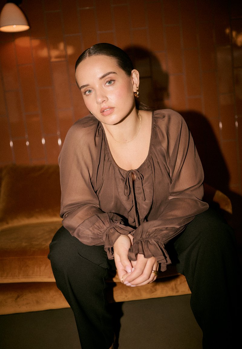 Blusa marrón transparente con lazo frontal y mangas con volantes. Combinada con pantalones negros. Modelo sentada en un sofá de terciopelo color beige sobre un fondo cálido.