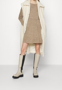 Beige Gummistiefel bis zum Knie mit schwarzen Akzenten, strukturiertem Obermaterial und klobiger schwarzer Sohle. Kombiniert mit einem hellbraunen Kleid und einem cremefarbenen ärmellosen Mantel.