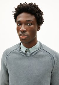 Grijze gebreide trui met geribde ronde hals, voorzien van een gladde textuur en verfijnde stiksels op de schouders, gelaagd over een lichtblauw overhemde.