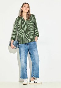 Camicia verde a maniche lunghe con strisce verticali nere, abbinata a jeans larghi blu chiaro e sneakers bianche. Tiene una pochette di denim.
