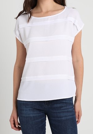 Kobieta w białej bluzce z krótkim rękawem z poziomymi teksturowanymi paskami i ciemnoniebieskich dżinsach na jednolitym tle.