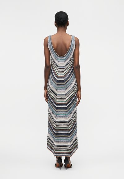 Moteris su tamsia oda ir surištais plaukais kukulyje, vilkinti rankovių neturinčią daugiaspalvių dryžių maxilango su gilia iškirpte nugaroje ir juodais aukštakulniais sandalais.