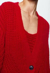 Punainen neulecardigan syvällä V-kaula-aukolla, teksturoidulla kuviolla ja yhdellä mustalla nappikoristeella. Kerroksittain mätsäävän punaisen topin päällä.