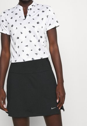 Persoon draagt een wit topje met korte mouwen met zwarte bloemenprints en een zwarte Nike-rok tegen een effen lichte achtergrond.
