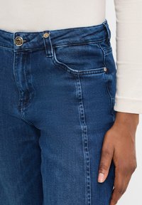 Dunkelblaue Jeans aus Denim mit glatter Textur, ausgestattet mit einer Fronttasche, goldenen Knöpfen und einer taillierten Passform.