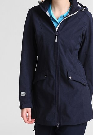 Kvinde iført en marineblå hættejakke med lynlås og klaplommer over en lyseblå skjorte med krave, stående foran en ensfarvet baggrund.