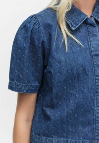 Denim short-sleeve shirt met een gestructureerd kruispatroon en puntige kraag, gedragen door een persoon met lang blond haar.