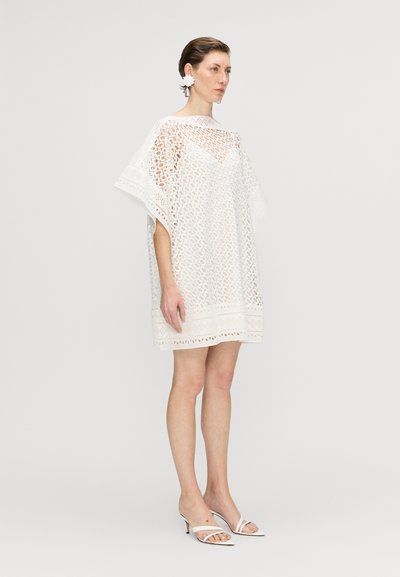Rochie albă din dantelă cu un croi lejer, prezentând un model geometric din dantelă și o margine ondulată. Asortată cu tocuri albe cu bretele.