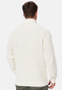 Pull blanc en tricot avec un col haut, des poignets côtelés et un motif texturé. Le dos présente une coupe ample et un design à manches raglan.