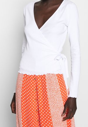 Top blanc à manches longues croisé avec un décolleté en V profond et un lien à la taille, associé à une jupe orange à motifs avec des pois et des sections florales.