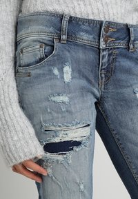 Jean en denim bleu clair usé avec des patchs effilochés, des accents bleu foncé, deux fermetures à bouton et des poches avant avec des détails de couture visibles.