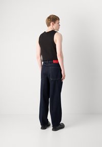 Ung mand står med ryggen til, iført en sort ærmeløs top, mørke vide jeans med et rødt "HUGO" mærke og sorte sko.