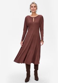 Robe longue marron en tissu doux, présentant une légère silhouette en A, un décolleté en forme de clé et des manches longues ; portée avec des bottes marron à hauteur de genou.