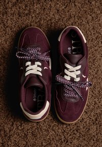 Bordeaux ruskinds-sneakers med cremefarvede accenter, teksturerede snørebånd med mønster og en brun gummisål. Topudsigt på et brunt tæppe.