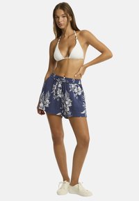 Kobieta w białym bikini i niebieskich szortach w kwiatowy wzór pozuje z pewnością siebie, nosząc białe trampki, na jednolitym białym tle.