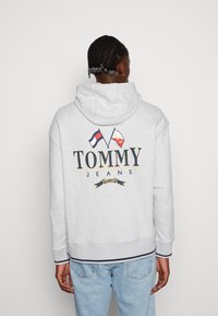 Felpa grigia chiara con cappuccio, caratterizzata da un grande logo stampato con bandiere e il testo "TOMMY JEANS". Realizzata in misto cotone con polsini e orlo a coste. Design casual.