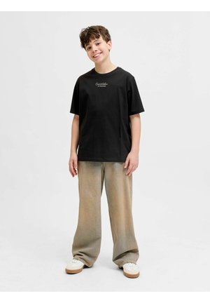 Ragazzo sorridente in piedi, indossa una maglietta nera a maniche corte, pantaloni beige a gamba larga e scarpe da ginnastica bianche su uno sfondo bianco semplice.