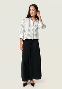Chemise blanche avec motif noir, coupe oversized et manches retroussées. Jupe maxi noire avec un détail noué à la taille. Escarpins noirs à bout pointu.