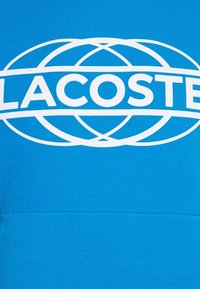 Blue sweatshirt featuring a large white circular logo with "LACOSTE" text in the center. Smooth fabric texture, casual design, hooded style.