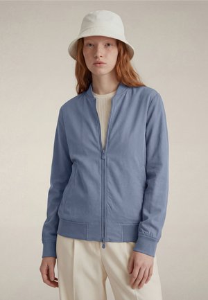 Młoda kobieta o rudych włosach nosi jasno-niebieską kurtkę na suwak, kremowe spodnie oraz biały kapelusz typu bucket, stojąc na jednolitym tle.