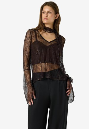 Žena má na sebe čierny priesvitný čipkovaný top s dlhým rukávom a detailom šatky a čierne nohavice, stojí s rukami pri vreckách, pozerá sa do strany.