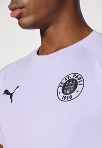 Camiseta lila con cuello redondo, que presenta un logo negro de Puma en el lado izquierdo y un emblema circular del FC St. Pauli en el pecho.