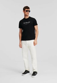 Camiseta negra con logo blanco, pantalones color crema y zapatillas negras. Gafas de sol con lentes oscuros. Diseño de atuendo simple y casual.
