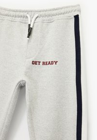 Pantalones de chándal grises con una textura suave, con un estampado en burdeos que dice "PREPÁRATE" y franjas laterales en azul marino, cinturilla elástica con cordón.