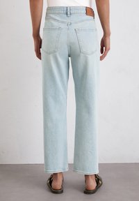 Persona vistiendo jeans de pierna recta azul claro y sandalias negras abiertas por detrás, de pie sobre un suelo gris frente a una pared blanca.