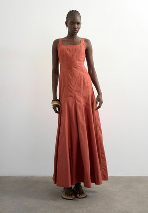 Vrouw in een mouwloze, vloerlange roestoranje jurk, blootsvoets staand tegen een witte muur en betonnen vloer.