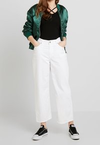 Chaqueta bomber de satén verde, top negro con tirantes entrecruzados, pantalones anchos blancos y zapatillas negras con suelas blancas.