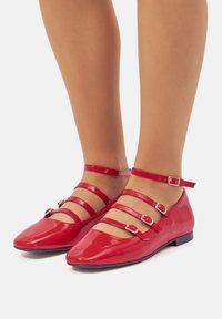 Bailarinas de charol rojo con punta afilada, que cuentan con tres correas en el tobillo con hebillas y un tacón bajo. Textura suave y acabado brillante.
