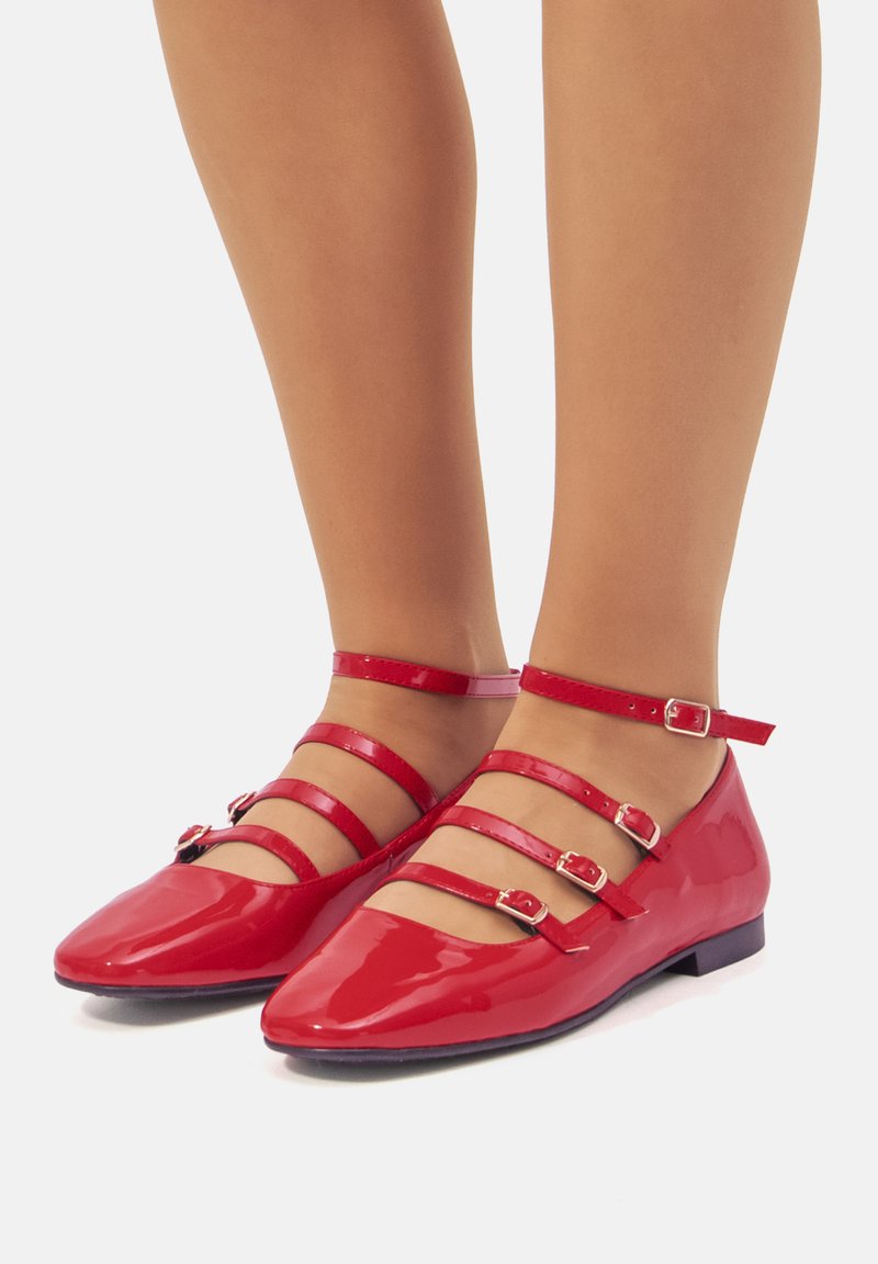 Bailarinas de charol rojo con punta afilada, que cuentan con tres correas en el tobillo con hebillas y un tacón bajo. Textura suave y acabado brillante.
