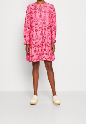 Vrouw die een loszittende roze jurk draagt met rode bloemenpatronen en lange mouwen, gecombineerd met crèmekleurige klompen met houten zolen.